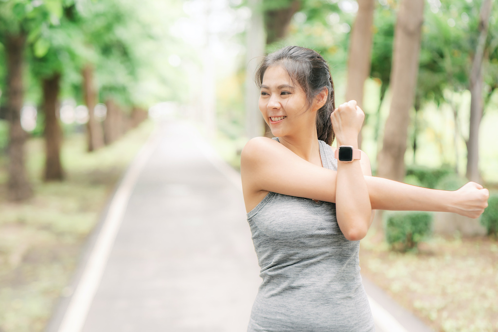 Young Asian woman with smartwatch doing some warm up exercises and shoulder streching before running at the park